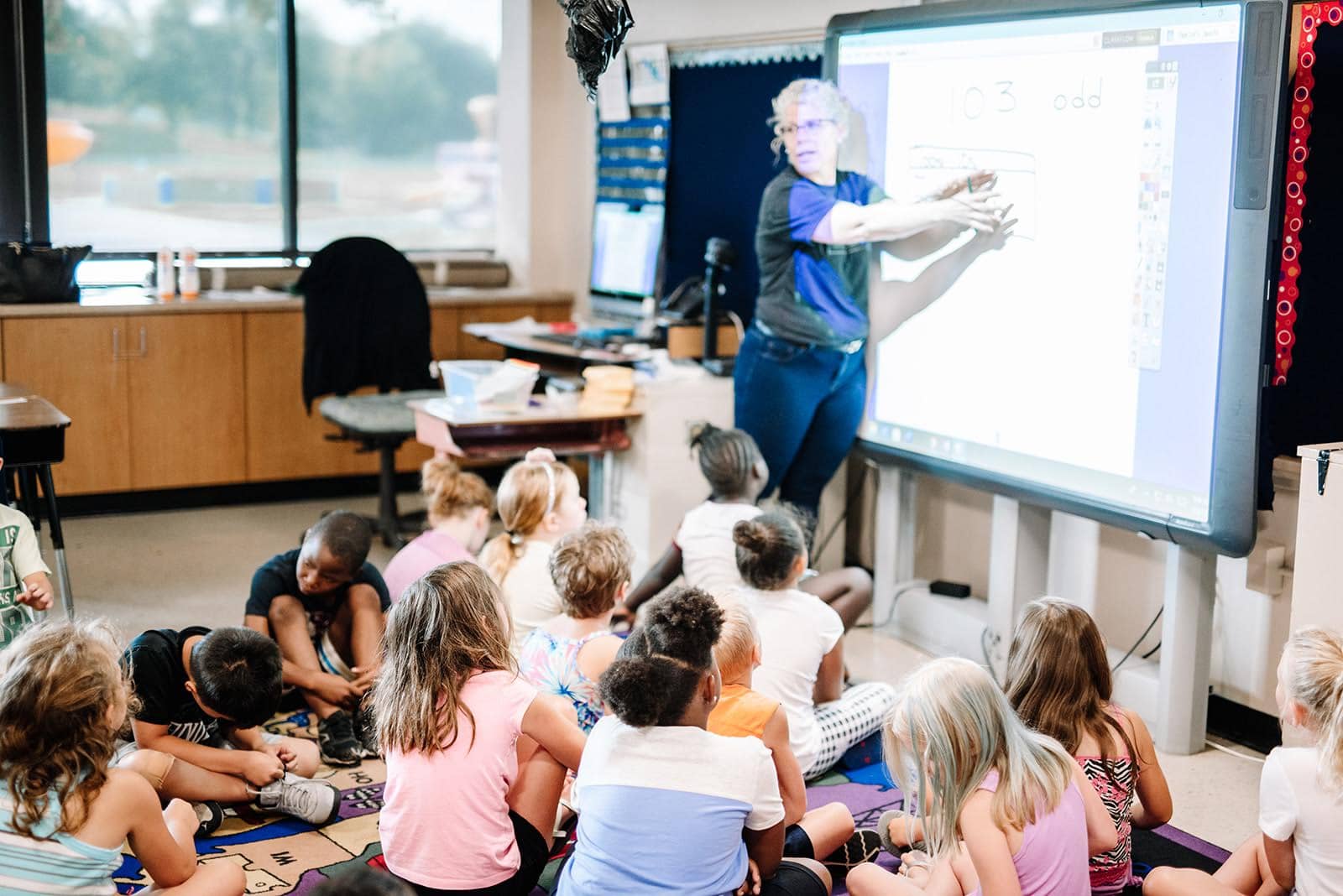 Photo of a teacher speaking to young students sitting on the floor.