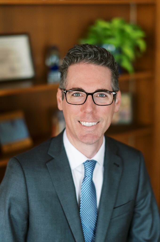 Professional photo of Joe Heitz, wearing a dark suit, white shirt, blue tie and glasses.
