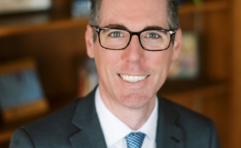 Professional photo of Joe Heitz, wearing a dark suit, white shirt, blue tie and glasses.